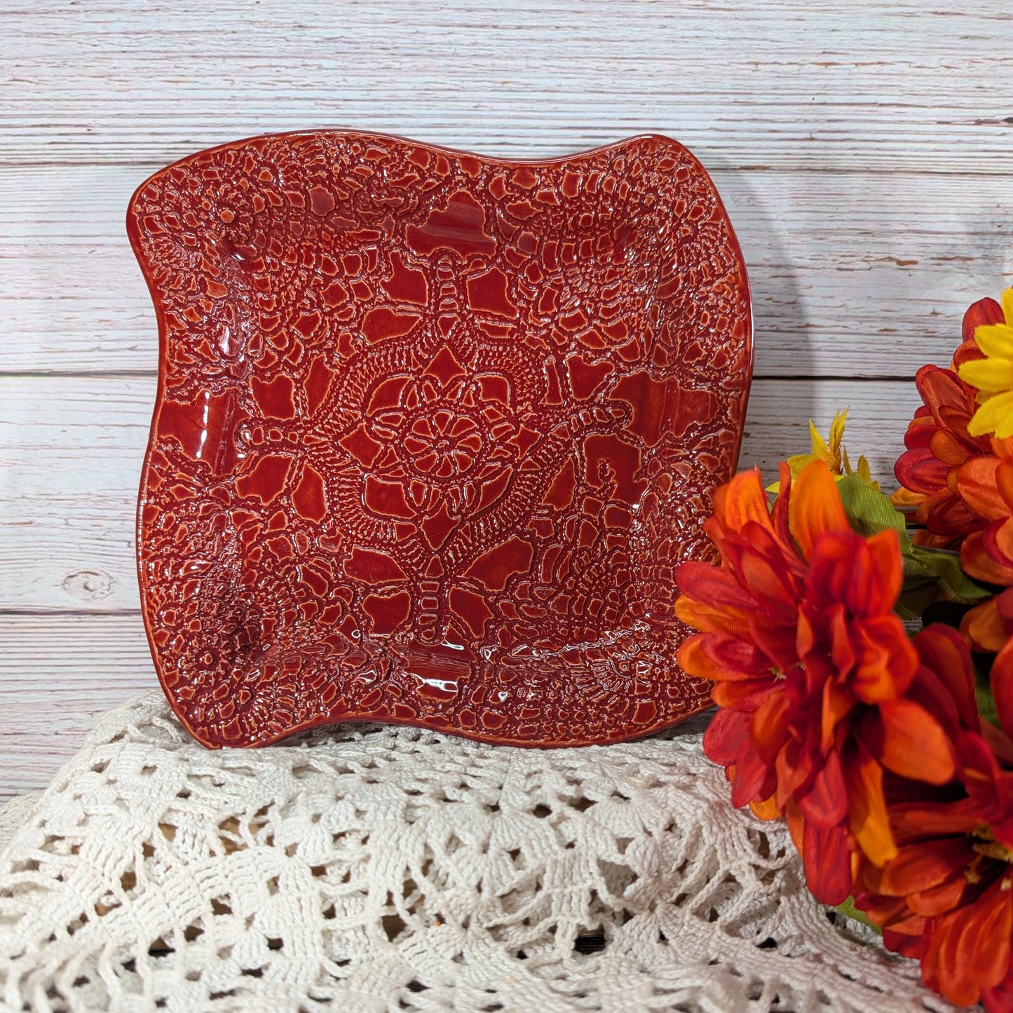 A square red ceramic plate with a raised floral lace pattern, styled on a white crocheted doily next to orange and yellow flowers against a white wood plank background.