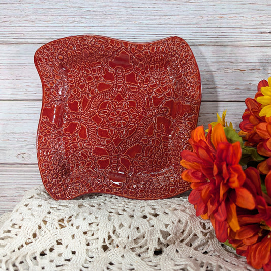 A square red ceramic plate with a raised floral lace pattern, styled on a white crocheted doily next to orange and yellow flowers against a white wood plank background.