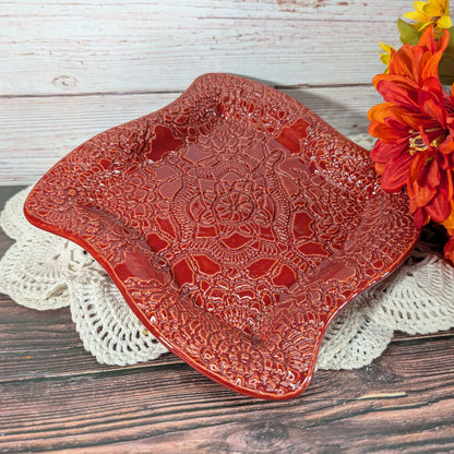 A glossy red square serving platter with a raised floral lace-like pattern, styled on a white crocheted doily over a rustic wooden table next to orange flowers.