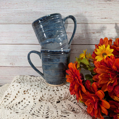 Two stacked dark blue-gray ceramic mugs with a textured, ribbed glaze, styled on a cream lace doily next to a bouquet of orange and yellow flowers against a whitewashed wood plank background.