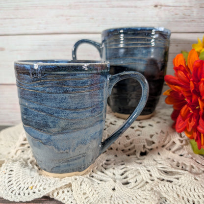 Two handmade ceramic mugs with a textured, swirled blue and gray glaze, displayed on a cream lace doily with a rustic white wood background and a red flower on the side.