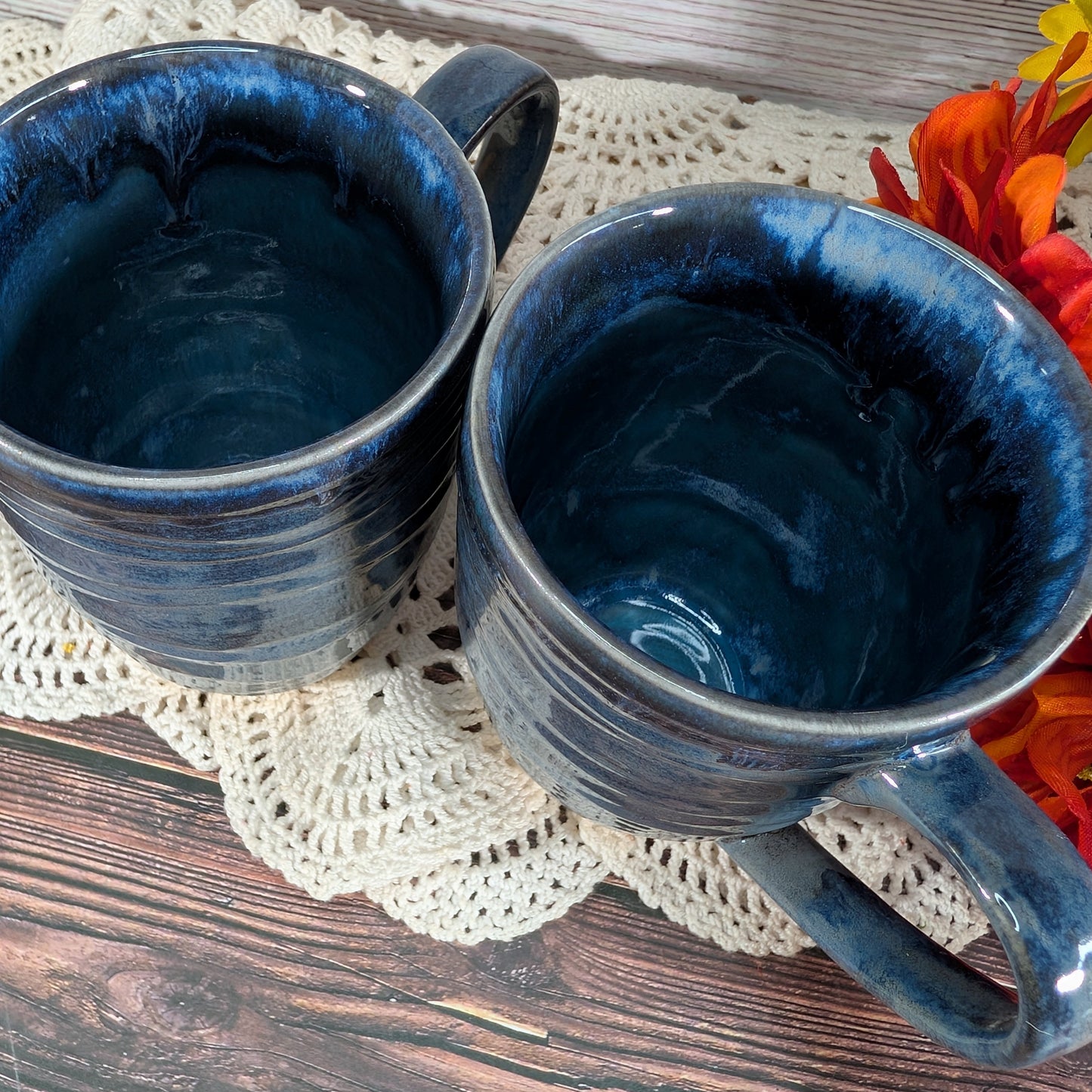 Two dark blue glazed ceramic mugs with a marbled finish, resting on a cream crocheted doily over a rustic wooden surface with orange flowers in the corner.
