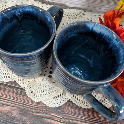 Two dark blue glazed ceramic mugs with a marbled finish, resting on a cream crocheted doily over a rustic wooden surface with orange flowers in the corner.