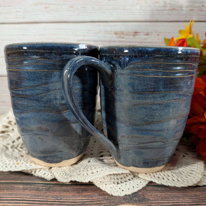 Two handmade ceramic mugs with a dark blue and gray wavy glaze, resting on a white lace doily over a wooden surface.