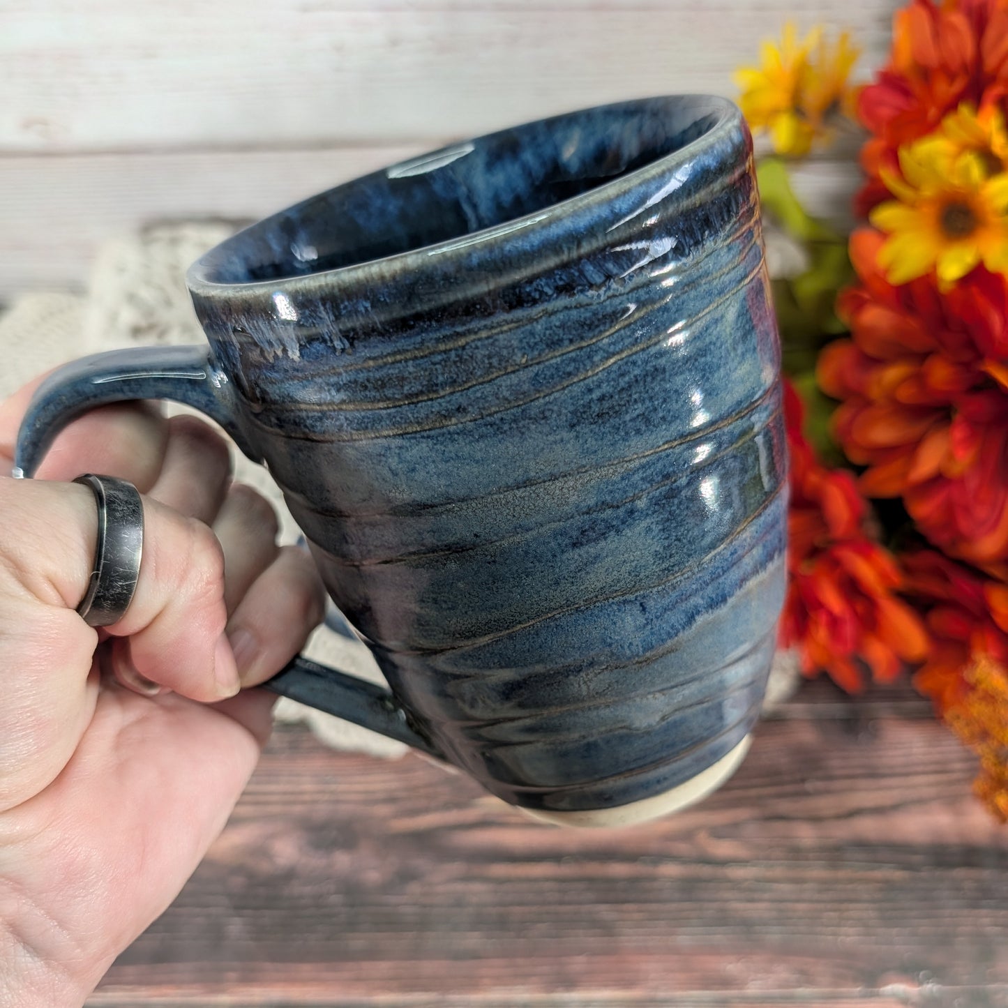 A hand holds a dark blue glazed ceramic mug with horizontal ridges, set against a rustic wooden background with orange and yellow flowers.