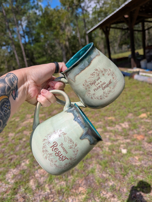 Two handmade ceramic mugs held up by a tattooed hand outdoors. The mugs have a light sage green glaze with dark brown dripping edges, a teal interior, and a brown floral wreath design with the words 'Together we can build something great' and 'Resist'.