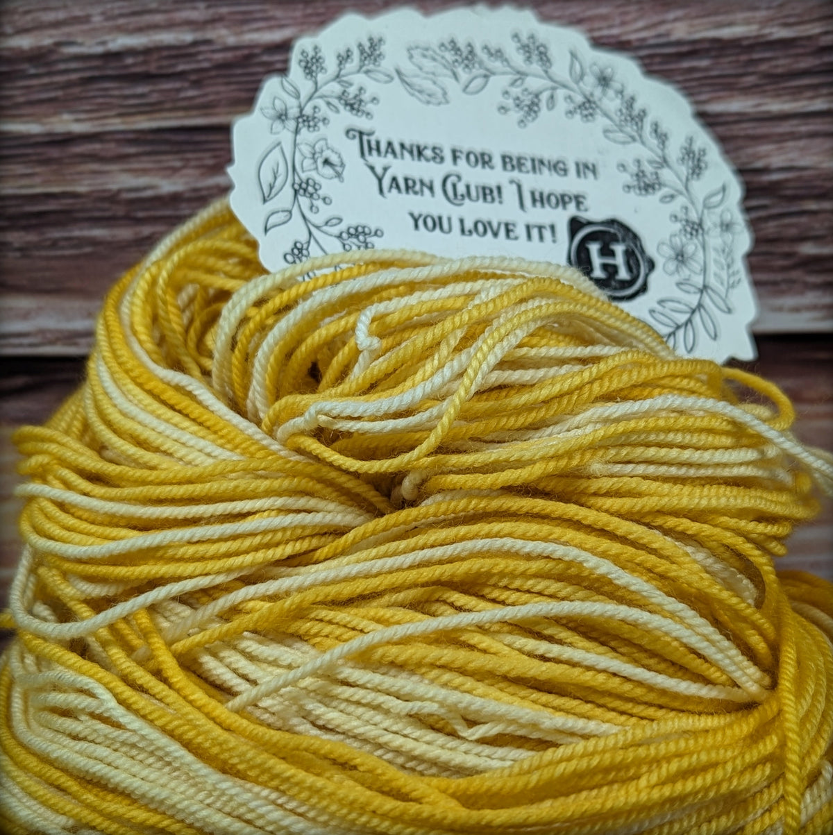 A skein of variegated yarn in shades of mustard yellow and cream, with a decorative paper tag that reads 'THANKS FOR BEING IN YARN CLUB! I HOPE YOU LOVE IT!' resting on a rustic wooden surface.
