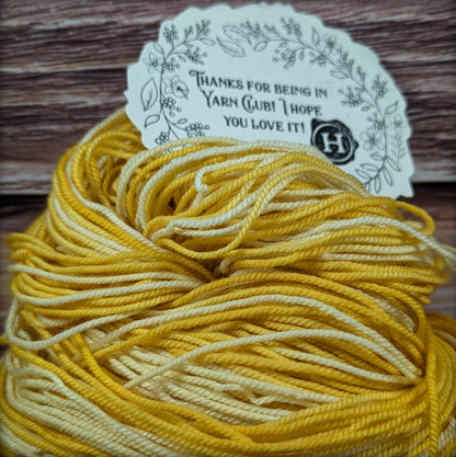 A skein of variegated yarn in shades of mustard yellow and cream, with a decorative paper tag that reads 'THANKS FOR BEING IN YARN CLUB! I HOPE YOU LOVE IT!' resting on a rustic wooden surface.