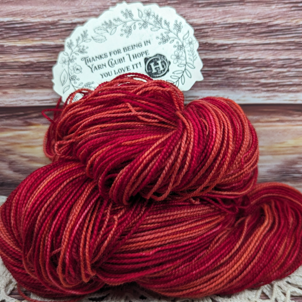 A skein of variegated yarn in shades of red, orange, and deep burgundy, with a white paper tag attached, set against a rustic wooden background.