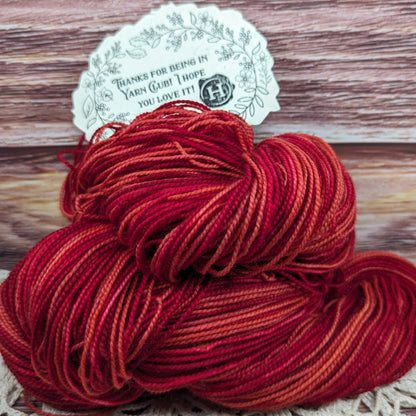 A skein of variegated yarn in shades of red, orange, and deep burgundy, with a white paper tag attached, set against a rustic wooden background.