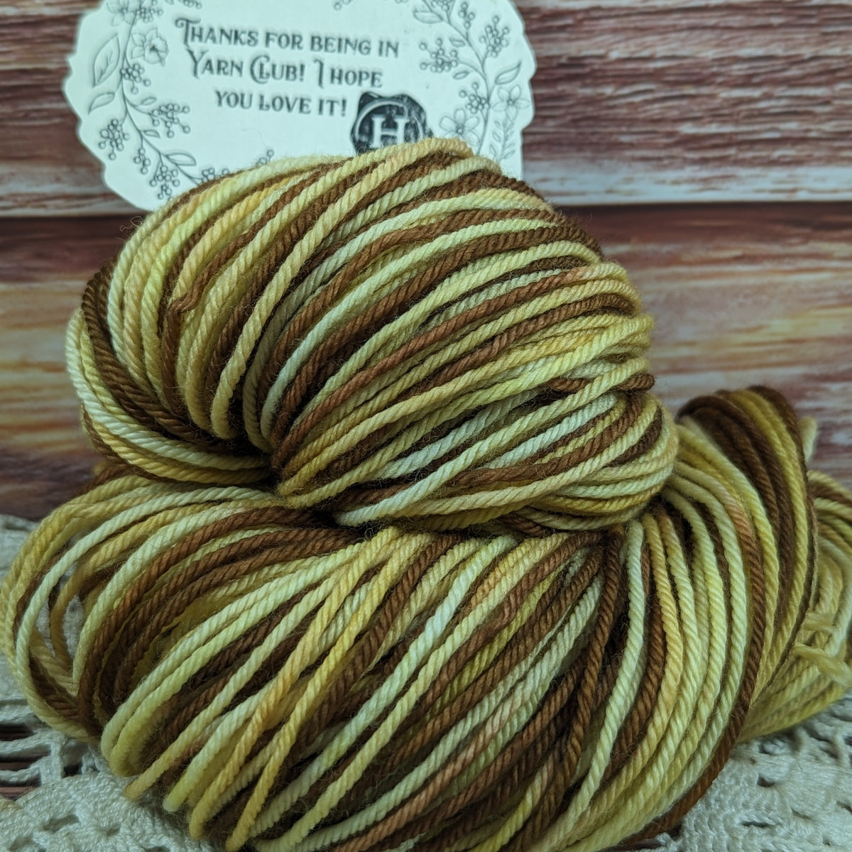 A skein of variegated yarn with alternating stripes of dark brown, medium brown, golden yellow, and cream, displayed on a lace doily with a wooden background and a paper tag.