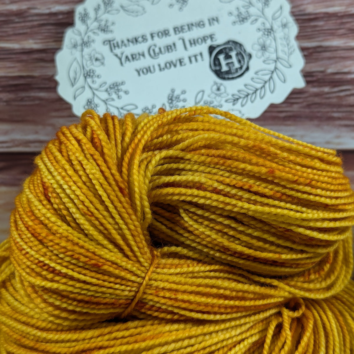 A skein of mustard yellow yarn with orange and brown variegated flecks, shown with a white paper tag that reads 'THANKS FOR BEING IN YARN CLUB! I HOPE YOU LOVE IT!' on a rustic wooden background.